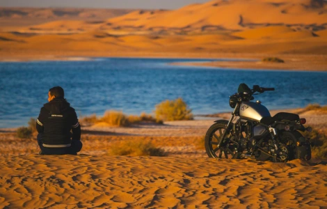 Motard contemplant un lac au milieu des dunes de sable au coucher du soleil.