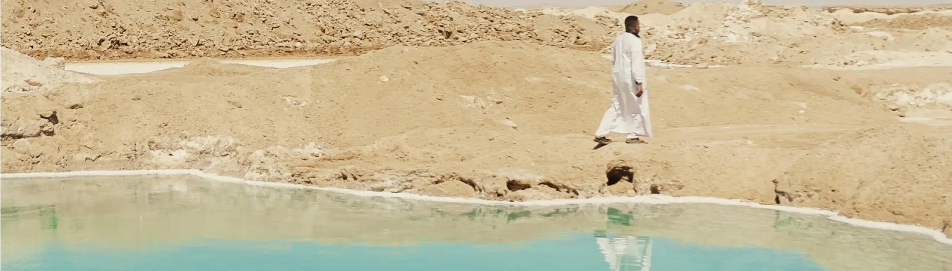 Paysage panoramique de l'Oasis de Siwa montrant un homme en habit traditionnel marchant au bord d'une source d'eau turquoise.