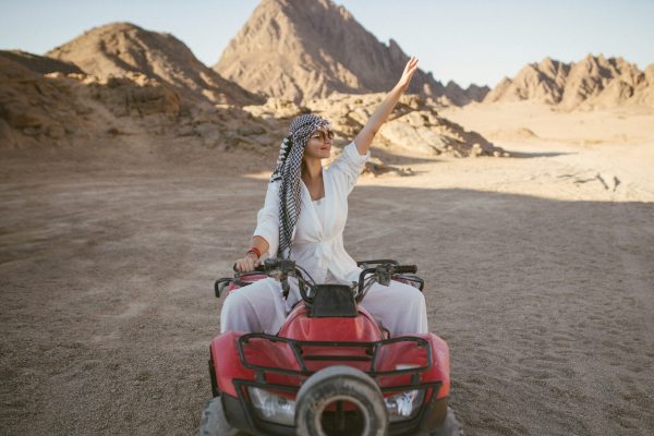 A rather attractive woman is going on a safari, driving an ATV in the desert. Beautiful young woman with a scarf on her head sits on a red four-wheeled ATV and rides towards adventure.