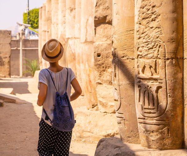 A young tourist in a white t-shirt and hat visiting the temple and looking at the ancient egyptian drawings on the columns of the Luxor Temple, Egypt